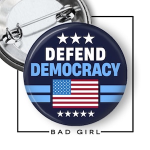 May include: A round, navy blue button with the words "DEFEND DEMOCRACY" in white and light blue. It features the American flag and the text "BAD GIRL". The button has a pin back.