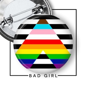 May include: A circular pin-back button featuring a horizontal black and white striped background overlaid with a large triangle containing horizontal bands of brown, light blue, pink, red, orange, yellow, green, blue, and purple, with the text BAD GIRL.