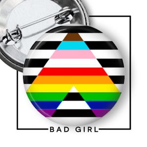May include: A round button pin with a black and white striped background and a rainbow triangle design. The pin has the text "BAD GIRL" in black letters. The pin is attached to a silver metal pin back.