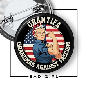 May include: A round black button with a graphic of a woman flexing her arm, set against an American flag background. Text on the button reads "GRANTIFA GRANDMAS AGAINST FASCISM" and "BAD GIRL".