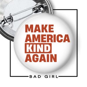 May include: A white button pin with the text "MAKE AMERICA KIND AGAIN" in red letters. The pin has a metal backing and a safety pin closure. The words "BAD GIRL" are at the bottom.