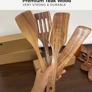 Może przedstawiać: Kolekcja drewnianych przybor&oacute;w kuchennych, w tym szpatułek i łyżek szczelinowych, wykonanych z wysokiej jakości drewna tekowego. Przybory mają ciepły brązowy kolor z widocznym usłojeniem drewna.