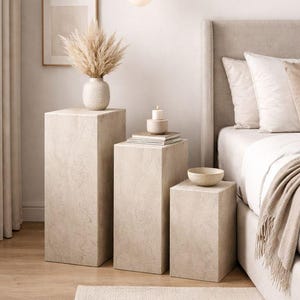 May include: A bedroom scene featuring three beige square pedestals of varying heights. The pedestals hold a vase with dried flowers, a stack of books, a candle, and a small bowl. A bed with neutral-toned bedding and a rug complete the scene.