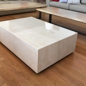 May include: A rectangular, cream-colored marble coffee table with a glossy finish. The table is a solid block design, sitting on a hardwood floor. The room also features a sofa and other tables.