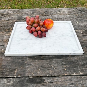 May include: A rectangular white marble serving tray with a slightly raised edge. A bunch of red grapes and a red and yellow apple rest on the tray, suggesting a food presentation theme.