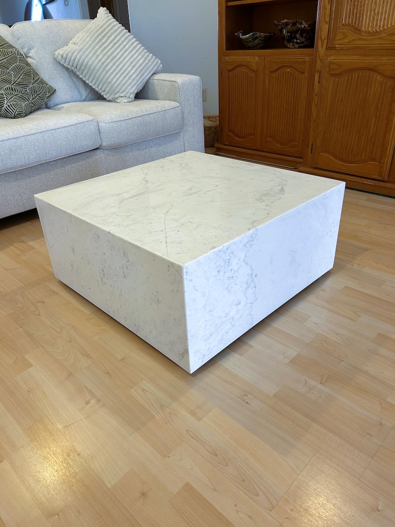 May include: A square, white marble coffee table with subtle gray veining. The table is a solid block shape, resting on a light wood floor. The table is in a living room setting, with a gray sofa and wooden cabinet in the background.