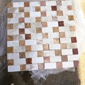 May include: A square mosaic tile sheet featuring a repeating pattern of rectangular and square stones in shades of cream, beige, tan, and reddish-brown. The natural stone pieces have varied textures and veining, arranged in a grid layout.