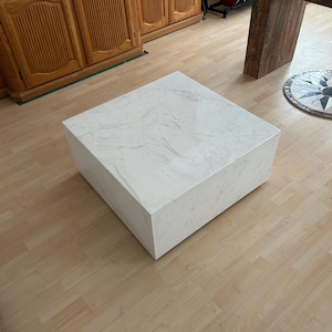 May include: A square, white marble coffee table with subtle gray veining. The table is a solid block shape, showcasing a minimalist design. The table is placed on a light wood floor, with wooden furniture and guitars in the background.