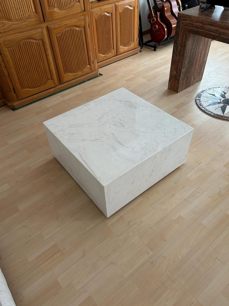 May include: A square, white marble coffee table with subtle gray veining. The table is a solid block shape, showcasing a minimalist design. The table is placed on a light wood floor, with wooden furniture and guitars in the background.
