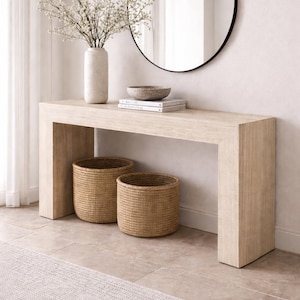 May include: A light wood-toned console table with two woven baskets below. A vase with white flowers, a stone bowl, and books sit on top. A round black-framed mirror hangs on the wall.