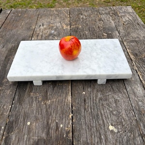 May include: A rectangular white marble serving board with a red and yellow apple on top. The board is supported by four small square feet. The apple is a mix of red and yellow colors.
