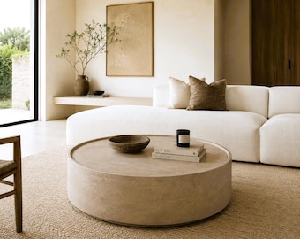 May include: A large, round, beige coffee table with a smooth, stone-like texture. On the surface sits a shallow, dark brown bowl, a stack of two books, and a small black candle in a cylindrical container, all arranged in the center.