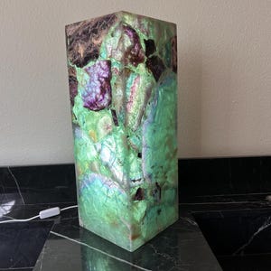 May include: A rectangular lamp with a mosaic design. The lamp features a mix of green, purple, and brown colors. The lamp is made of a translucent material and has a white power cord.