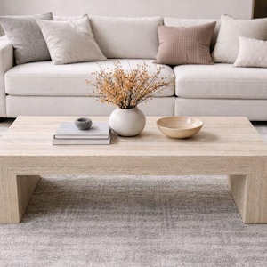 May include: A light-colored wooden coffee table with a rectangular shape and block legs. A white sofa with neutral-colored pillows sits behind the table. A white vase with dried flowers, a bowl, and books are on the table.