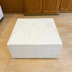 May include: A square, white coffee table with a marble-like pattern. The table is a solid block shape and sits on a light-colored wooden floor. The table is approximately 36 inches wide and 16 inches tall.