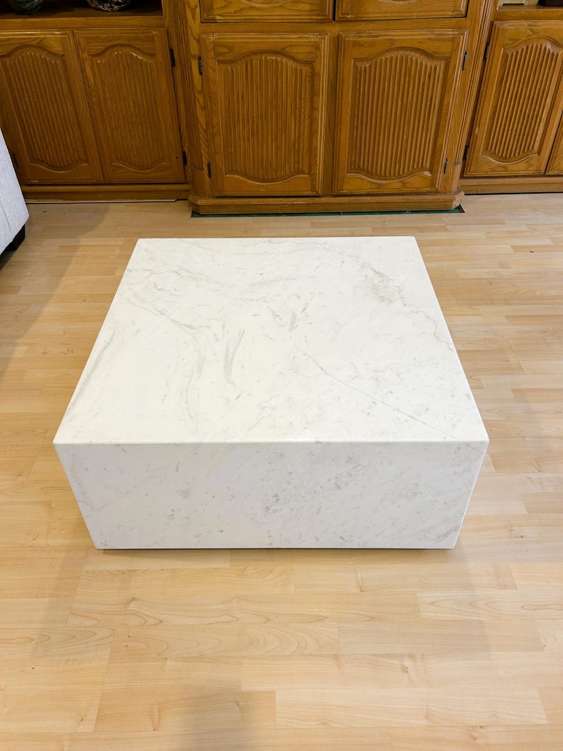 May include: A square, white coffee table with a marble-like pattern. The table is a solid block shape and sits on a light-colored wooden floor. The table is approximately 36 inches wide and 16 inches tall.