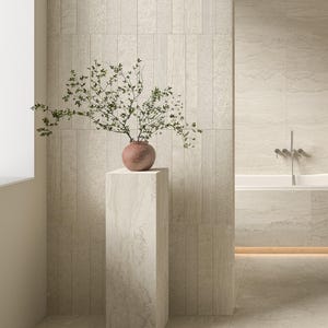 May include: A minimalist bathroom scene featuring a beige color palette. A terracotta vase with green branches sits atop a stone pedestal. The background includes textured wall tiles and a bathtub with chrome fixtures.