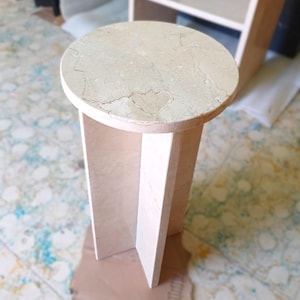 May include: A beige marble side table with a round top and a geometric base. The table is approximately 20 inches tall and 12 inches in diameter, showcasing natural veining patterns. The design is minimalist and modern.