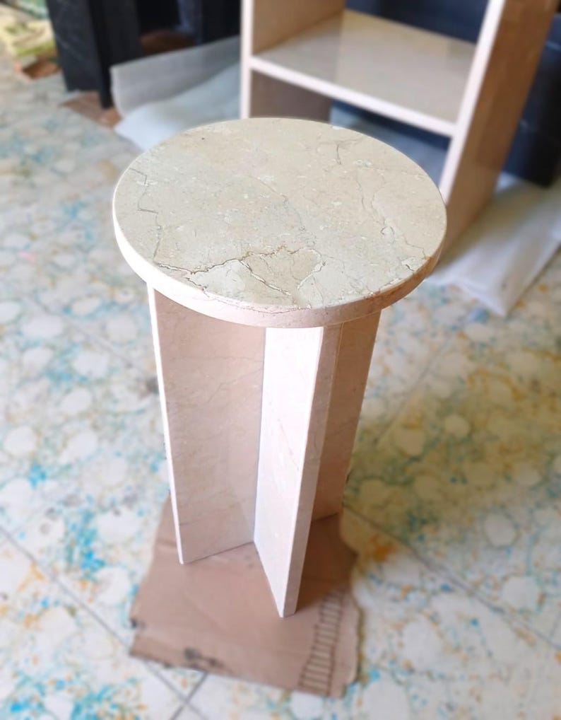 May include: A beige marble side table with a round top and a geometric base. The table is approximately 20 inches tall and 12 inches in diameter, showcasing natural veining patterns. The design is minimalist and modern.