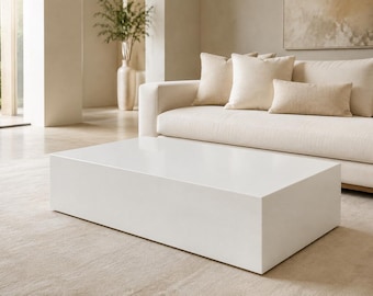 May include: A low-profile, rectangular coffee table with a smooth, solid white finish. The table features a clean, block-like structure with sharp edges and a flat surface, positioned horizontally in front of a light-colored sofa.