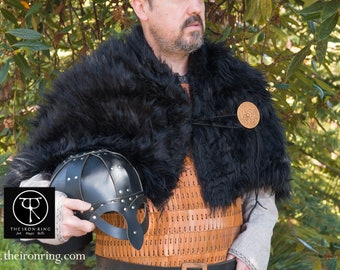 May include: A person wearing a black fur cloak with a leather and metal chest piece and a round metal brooch. The person is holding a black leather helmet with metal accents.