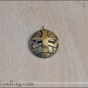 May include: A round, gold-colored pendant with a geometric design. The pendant has a raised, textured surface and a silver loop at the top.