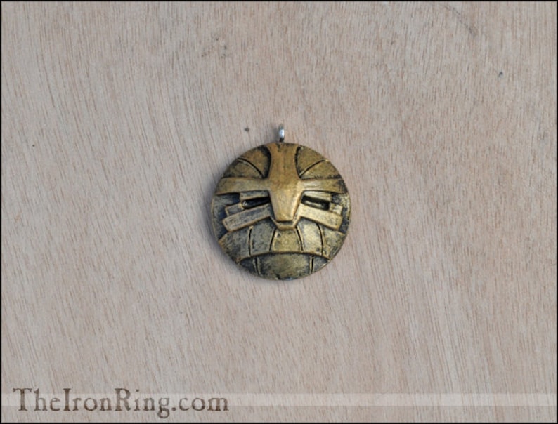 May include: A round, gold-colored pendant with a geometric design. The pendant has a raised, textured surface and a silver loop at the top.