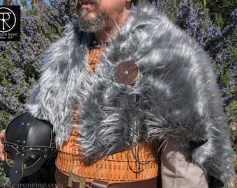May include: A person wearing a grey fur cloak with a leather strap and a round metal buckle. The person is also wearing a brown leather armor with a belt and a black helmet. The background is a green bush with purple flowers.