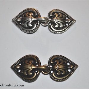May include: Two heart-shaped metal clasps, one silver and one gold, with intricate floral designs. The clasps are designed to be used as a closure for garments or accessories.