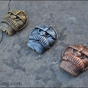 May include: Three metal pendants in gold, silver, and copper tones, each featuring a detailed, stylized owl design. The pendants are displayed on a textured, gray surface, showcasing their intricate craftsmanship.
