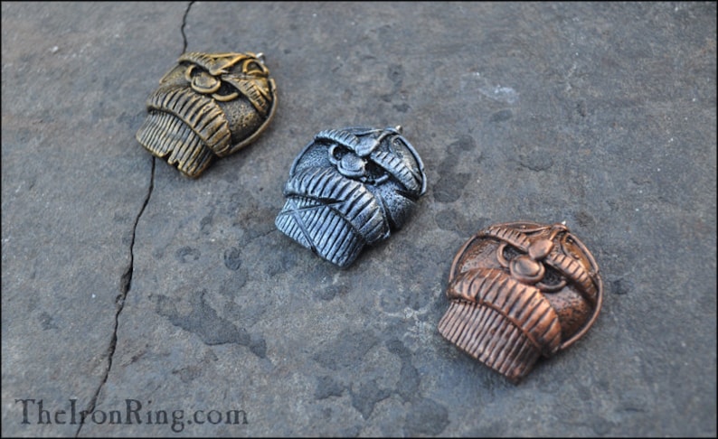 May include: Three metal pendants in gold, silver, and copper tones, each featuring a detailed, stylized owl design. The pendants are displayed on a textured, gray surface, showcasing their intricate craftsmanship.