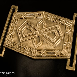 May include: A gold metal belt buckle with an intricate geometric design. The buckle has two loops on either side for attaching a belt.