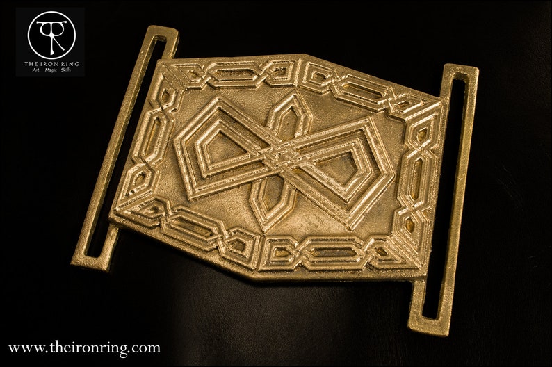 May include: A gold metal belt buckle with an intricate geometric design. The buckle has two loops on either side for attaching a belt.