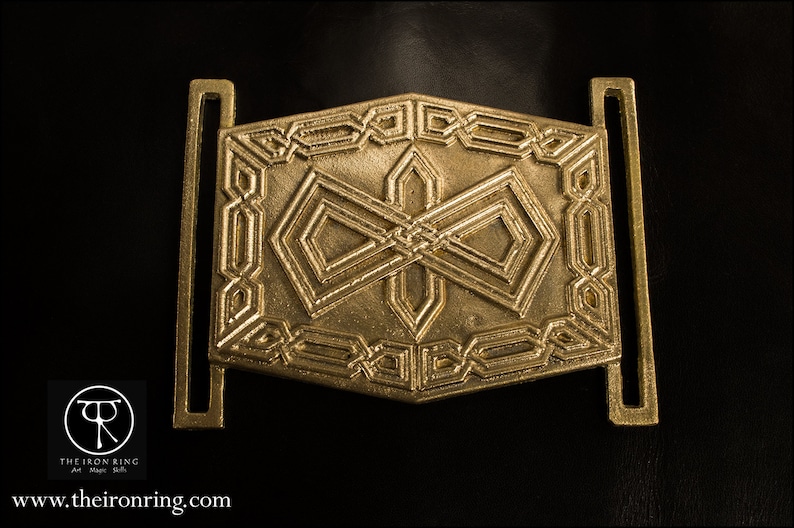 May include: A gold-colored metal belt buckle with an intricate geometric design. The buckle has a square shape with rounded corners and features a repeating pattern of interlocking shapes.