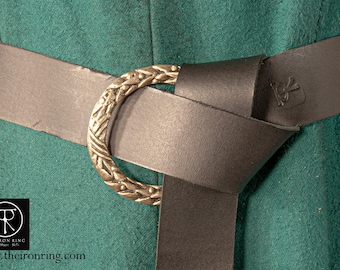May include: A close-up of a brown leather belt with a decorative brass buckle. The buckle is ornate and features a detailed design. The belt is tied in a knot and the buckle is secured.