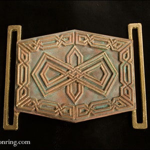May include: A decorative metal belt buckle with a geometric design in shades of copper and green. The buckle has a rectangular shape with two loops on either side.