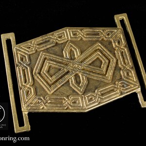 May include: A gold-toned metal belt buckle with an intricate geometric design. The buckle features a repeating pattern of squares and diamonds.