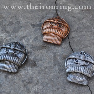 May include: Three metal decorative pendants with a detailed, stylized design. One is bronze, and two are silver. The pendants are arranged on a textured gray surface. The website address "www.theironring.com" is visible in the top left corner.