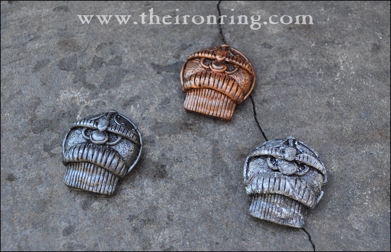 May include: Three metal decorative pendants with a detailed, stylized design. One is bronze, and two are silver. The pendants are arranged on a textured gray surface. The website address "www.theironring.com" is visible in the top left corner.
