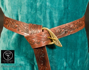 May include: Brown leather belt with an embossed floral design and a brass buckle. The belt is wrapped around a teal garment. The Iron Ring logo is visible in the lower left corner.