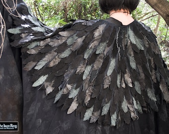 May include: A black cape with layered feather detailing. The feathers are primarily black with accents of gray and pale green. The cape is worn over the shoulders, with the back of the neck visible. The Iron Ring logo is in the lower left corner.