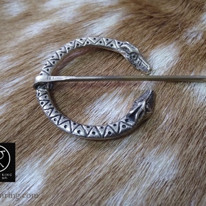 May include: Silver circular brooch with two dragon heads on either side of the pin. The brooch is decorated with a geometric pattern of triangles.