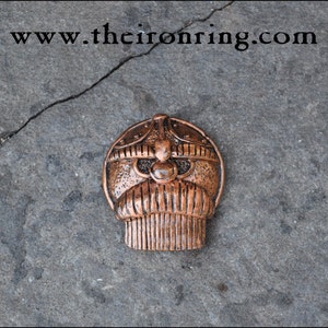 May include: A copper-colored metal pendant with a stylized face design. The pendant has a rounded top with a detailed helmet-like structure and a beard-like bottom. The website address "www.theironring.com" is visible at the top.