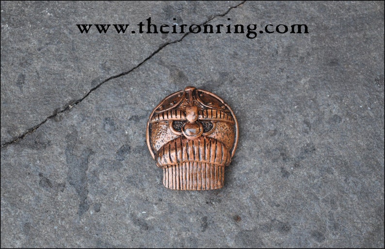 May include: A copper-colored metal pendant with a stylized face design. The pendant has a rounded top with a detailed helmet-like structure and a beard-like bottom. The website address "www.theironring.com" is visible at the top.