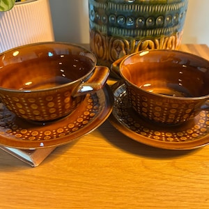 May include: Two brown ceramic soup bowls with matching saucers, each featuring a glossy finish and a repeating embossed dot pattern. The bowls have two small side handles and a deep, rounded shape, while the saucers have a wide rim.