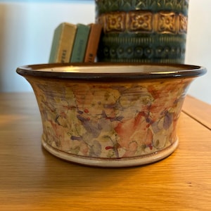 May include: A ceramic bowl with a wide, flared rim. The bowl has a colourful, abstract design with shades of orange, purple, and yellow. The bowl is on a wooden surface. A tall, green and yellow vase is in the background.