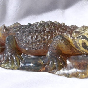 Puede incluir: Una escultura detallada de un cocodrilo en ámbar, de color marrón amarillento y con detalles intrincados. El cocodrilo está acostado sobre su vientre con la cabeza girada hacia un lado.