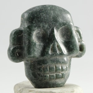 May include: A carved dark green skull sculpture with detailed eye sockets, nose, and teeth. The skull is made of a stone-like material and sits on a light-colored base. The sculpture has a weathered appearance.