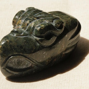 May include: A dark green carved stone sculpture of a stylized animal head. The sculpture has detailed features, including eyes, ears, and textured surfaces. The stone has a mottled appearance with variations in color.