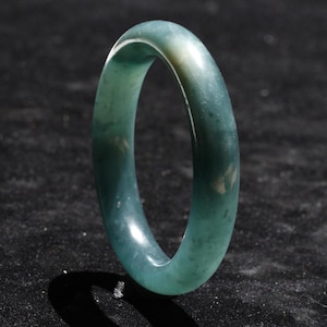 May include: A jade bangle bracelet in shades of teal and green. The smooth, polished surface reflects light, highlighting the natural variations in color. The circular design is a classic jewelry style.
